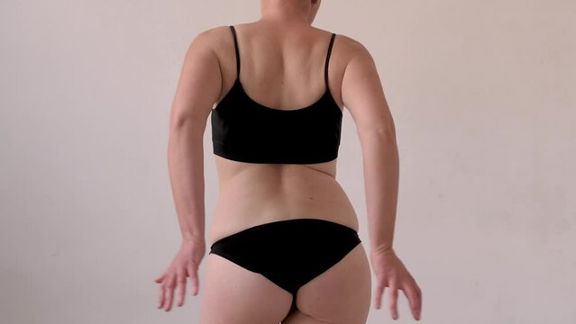 A woman in black lingerie sorts out the folds on her stomach after losing weight and dieting, dancing and twirling her hips. Waist creases and cellulite. Overweight and weight loss. View from the back