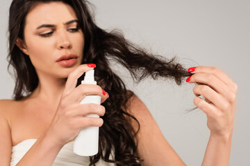 brunette woman spraying treatment product on damaged hair isolated on grey.