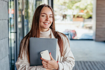 student holding a smartphone and a folder looking at camera against the background of an office...