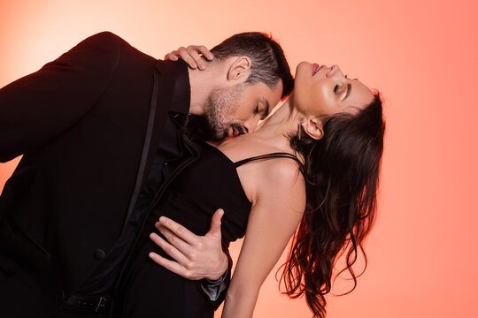 passionate and bearded man kissing chest of sexy woman in black dress on pink.