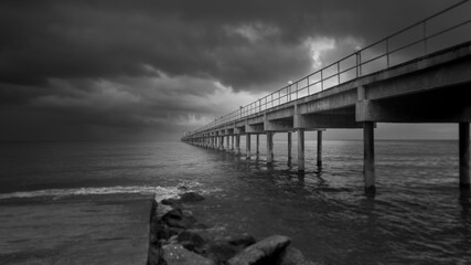 Long exposure fine art black and white image of a long pier into the ocean. Silky smooth sea water washes around the concrete pillar. Dark exposure reinforces the gloomy impression. Copy space concept