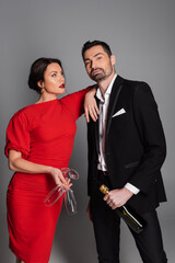 Sexy woman in red dress holding glasses and looking at boyfriend with champagne on grey background.