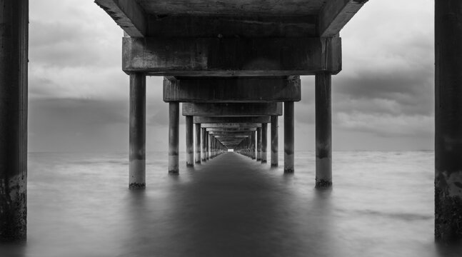 Long exposure fine art black and white image of a long pier into the ocean. Silky smooth sea water washes around the concrete pillar. Dark exposure reinforces the gloomy impression. Copy space concept
