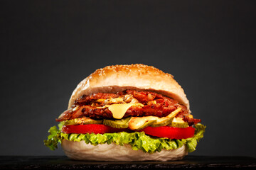 Burger with bacon onions tomatoes cucumber lettuce and french fries against dark gray background.