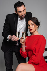 Elegant man in suit holding champagne while girlfriend in dress looking at camera on grey background.
