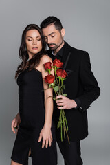 bearded man in suit holding red roses near brunette woman isolated on grey.