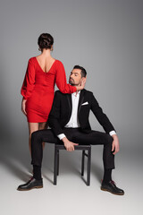 Elegant woman in dress touching face of boyfriend sitting on chair on grey background.