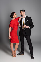 Elegant man in suit holding champagne bottle and glasses near girlfriend on grey background.