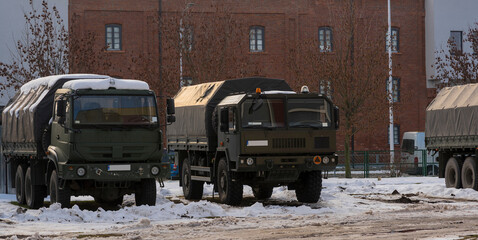 Wojskowe ciężarówki terenowe , polskiej produkcji zaparkowane na zaśnieżonym placu , na tle budynku z czerwonych cegieł .