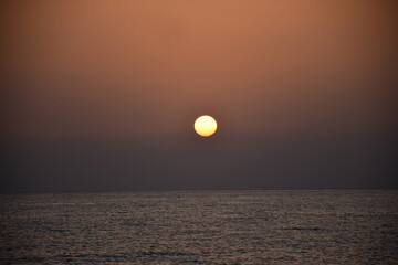 The sun rises over the sea from behind the mountains. ripples on the surface of the water. Dawn in dark colors. Round sun without rays