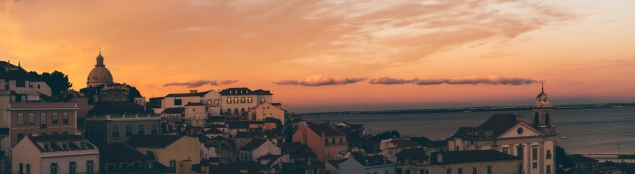 A panoramic image with a cityscape of an antique part of Lisbon with old houses, churches, and a river with a stunning sunset in the background; a panorama of an old Portuguese city, dramatic evening