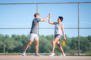Laughing couple playing pickleball game, hitting pickleball yellow ball with paddle, outdoor sport leisure activity, celebrating victory .