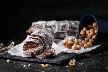 Chocolate salami filled with almonds, hazelnuts and raisins on the black background. Italian sweet sausage shaped dessert sprinkled with powdered sugar. Christmas and New Year festive pastry