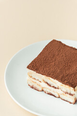 Homemade tiramisu cake on white plate against beige background. Traditional Italian no-bake dessert made of savoiardi, filled with mascarpone cheese, coffee espresso and sprinkled with cocoa powder