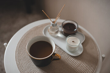 Cup of fresh coffee with scented candles and liquid home fragrance in glass bottle with bamboo sticks on table in room closeup.