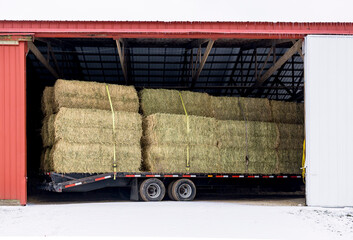 A double axle trailer loaded with big square bales of hay in a red pole shed with snow on the...