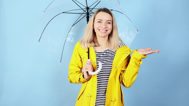 Portrait of happy young blond girl looking around enjoying rain day under umbrella and try to catch drops on isolated blue background Smiling female in yellow raincoat looking at the camera at studio