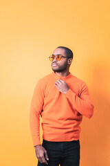 Obraz premium A dark-skinned man posing in front of an orange wall wearing orange sunglasses, the boy looks to the side as he readjusts his orange sweater.