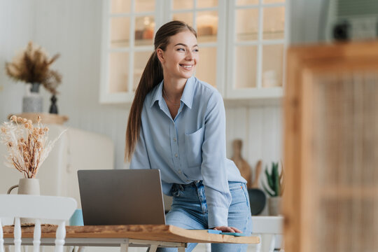 Gorgeous blonde young woman in blue shirt and blue jeans standing at desk with laptop looks aside toothy smiles against blurry kitchen home. Pretty female model with ponytail remote working home.