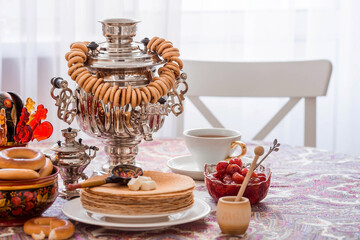 Russian traditions. Russian holiday Maslenitsa. Still life with a cup of tea, a stack of pancakes, sour cream, honey, cubes of butter, bagels, dried fruits, caramel cockerel and samovars.