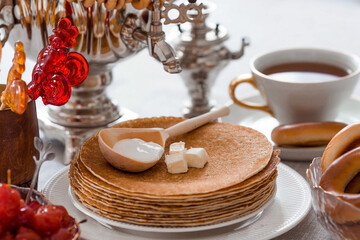 Russian traditions. Russian holiday Maslenitsa. Still life with a cup of tea, a stack of pancakes, sour cream, honey, cubes of butter, bagels, dried fruits, caramel cockerel and samovars.