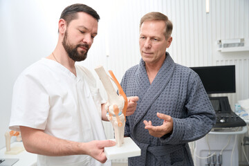 Professional doctor showinf knee joint replica to middle-aged man