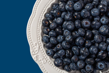 Blueberries on a white plate over blue background closeup. Ripe wild berries for healty vegan snack. Sweet blueberry as vitamin vegetarian dessert. Natural antioxidant. Detox diet.