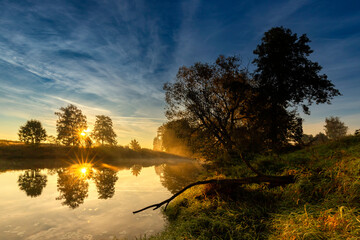 Wschód słońca. Jesienny poranek nad dziką rzeką w Polsce. Odbicia w wodzie. Wodny krajobraz. Świt. Kolory jesieni. Jesienne scenerie. Mgliste poranki. Bez ludzi. Tło natury. © anettastar