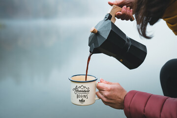 Young woman hiker pouring fresh coffee into cup or mug from moka pot coffee maker outdoor. Tourism...