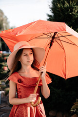 A little girl with loose hair in a hat and a red dress with a big red umbrella outside in clear weather