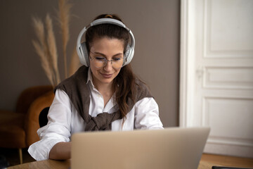 A calm young brunette with glasses, a woman in a white shirt listens to music or an audiobook with headphones and works on a laptop. The concept of people's lifestyle.