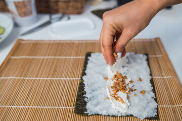 Unrecognizable woman's hand pouring crispy onion on top of cheese spread on a sheet of nori seaweed for ready to make homemade sushi at her kitchen table.