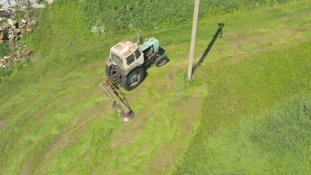 Old tractor carefully mows the grass around a concrete pole. Summer Haymaking with a tractor mower. Tractor mows green grass to dry hay - top view overhead aerial shot. 