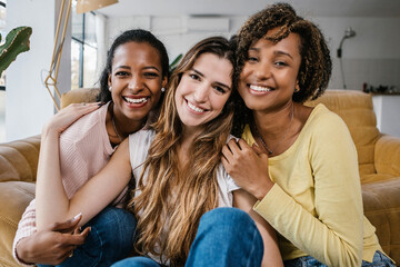 Portrait of united multiracial female friends smiling at camera at home interior. Happy mixed race...