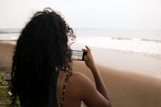 Rear view of woman photographing while standing at beach against sky