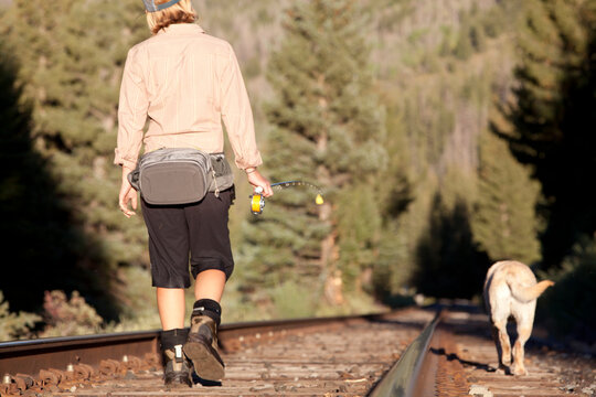A woman and her dog walk along some train tracks to their next fishing spot.