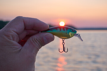 Male fisherman holding a small wobbler lure with a scenic sunset on a lake on the background.