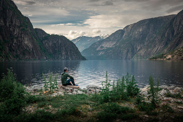 Fototapeta na wymiar Man sitting at waterfront of lake in the mountain landscape Eidfjord in Norway, looking into the fjord, clouds in the sky