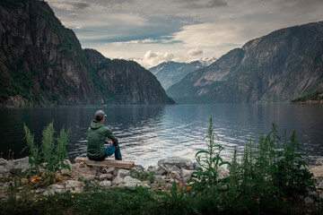 Man sitting at waterfront of lake in the mountain landscape Eidfjord in Norway, looking into the fjord, clouds in the sky