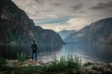 Fototapeta na wymiar Man standing at waterfront of lake in the mountain landscape Eidfjord in Norway, looking into the fjord, clouds in the sky