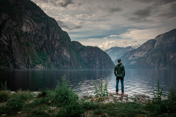 Man standing at waterfront of lake in the mountain landscape Eidfjord in Norway, looking into the fjord, clouds in the sky - 559031861