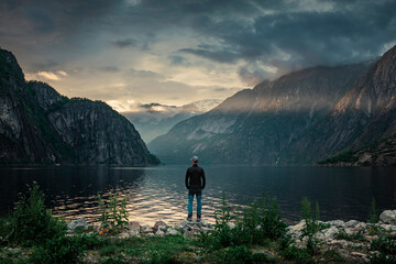 Man standing at waterfront of lake in the mountain landscape Eidfjord in Norway, looking into the fjord, clouds in the sky during sunset with sun beams