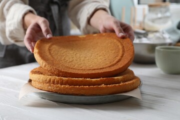 Woman stacking homemade sponge cakes at white wooden table in kitchen, closeup