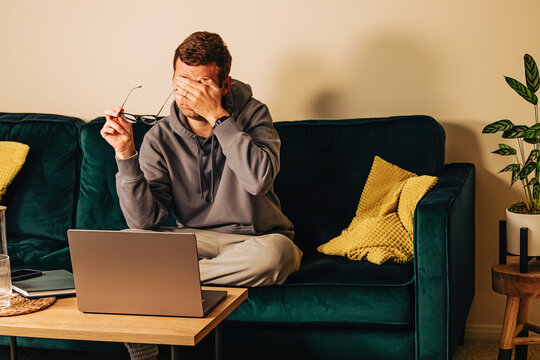 Sad, Frustrated Man Sitting On The Green Couch In His Living Room In Front Of His Laptop