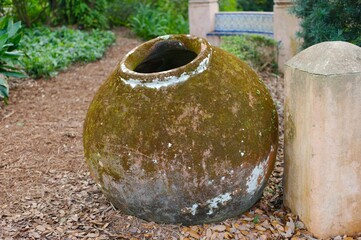 Old round water pot brown outdoors on ground cistern on brown path beside building. Old clay jar with a circular opening.