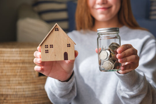 Closeup image of a woman holding wooden house models and a glass jar of coins for saving money concept