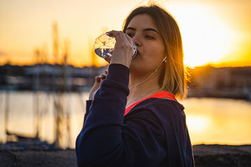 Young beautiful curvy woman taking a break from workout routine drinking some water from a plastic bottle at sunset