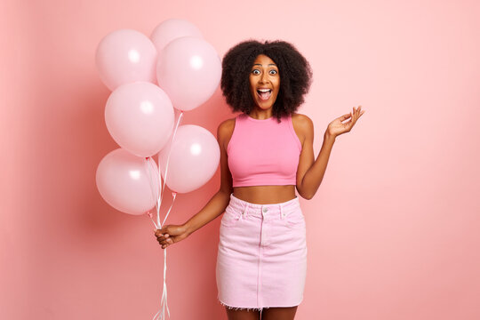 A Shot Of Positive Smile, Woman Holds Pink Balloons, Spread Hand A Side In A Huge Surprise, Opened Eyes And Mouth From Amazing, Isolated Next To Pink Wall