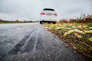 Close up of car brake path on edge of asphalt road on the background of parked white car, fallen yellow leaves on the grass and gray sky. Emergency braking tracks on edge of highway. - obrazy, fototapety, plakaty