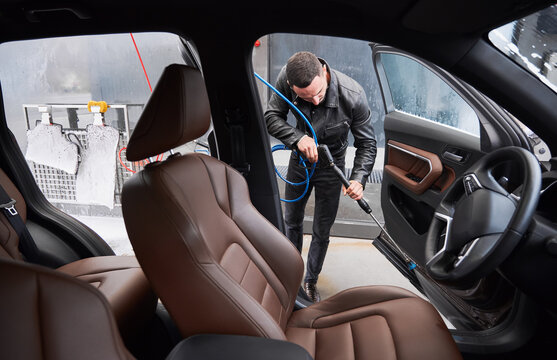 View From Inside A Car On Young Man Washing Car On Carwash Station Outdoor. Handsome Worker Cleaning Automobile, Using High Pressure Water.